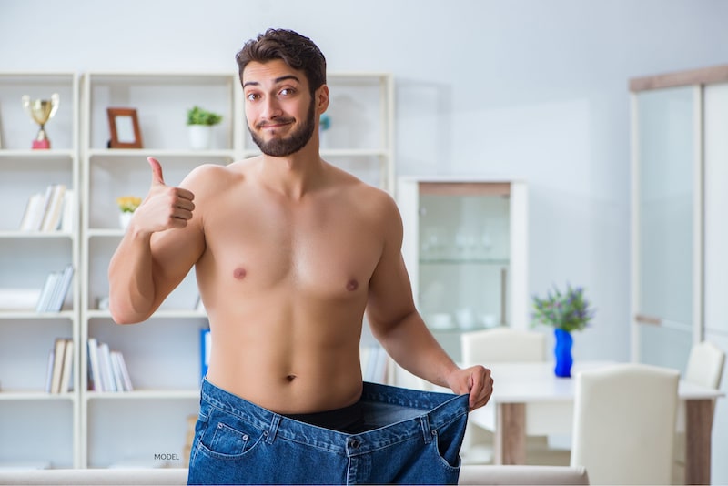 Attractive man who is shirtless and holding up an overly large pair of jeans while he gives a thumbs up